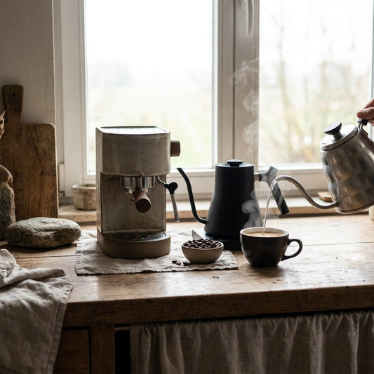 Barista-style home coffee scene with a fresh americano in a ceramic cup, espresso machine and kettle on a warm Belgian kitchen counter.